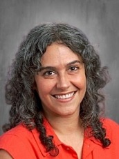 Professional head-and-shoulders portrait of a smiling woman with dark, curly hair in a black blazer and white blouse, against a neutral gray background.