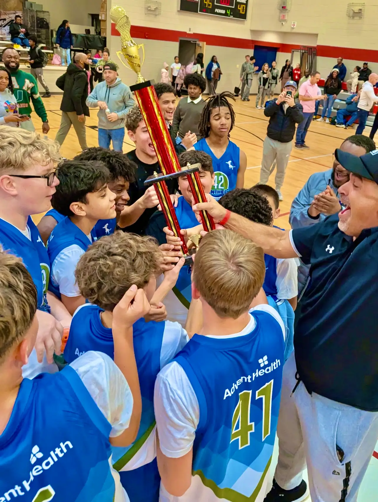 Forest Lake Education Center basketball team in blue and white jerseys hoisting a championship trophy with their cheering coach in a gymnasium