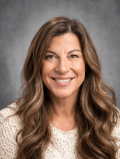Professional headshot: smiling woman with brown hair, wearing a black blazer and pink blouse, gray background.