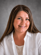Smiling woman with long brown hair wearing a white blouse, neutral studio background (headshot].)