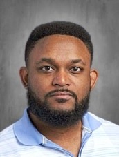Portrait of a man with short hair and a full beard, wearing a light blue polo shirt, facing the camera against a gray backdrop.